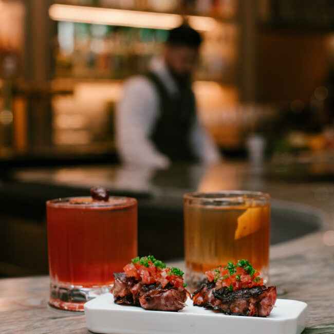 Hot capicola spiedini with diced tomato topping, served with a passion fruit old fashioned and classic old fashioned at Nic + Junior’s bar in River North