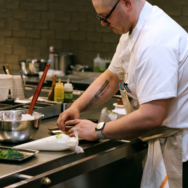 Chef Justin plating a dish at the kitchen pass at Nic + Junior’s, wearing a white coat and apron