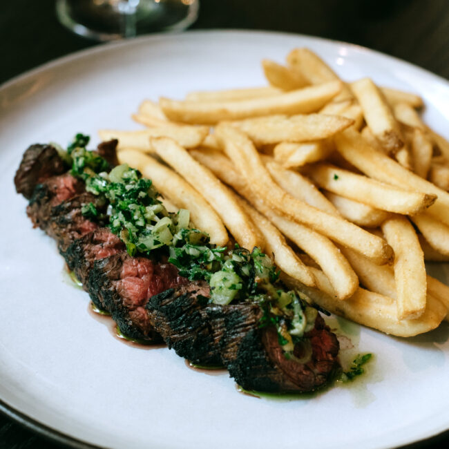 Charred bavette steak topped with herb sauce, served with golden frites on a white plate at Nic + Junior’s