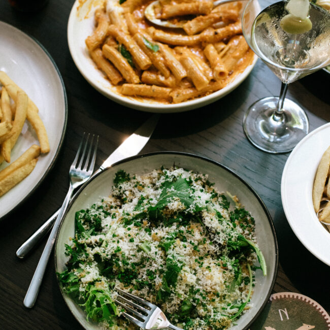 Overhead view of à la carte dishes at Nic + Junior’s, including rigatoni, caesar salad, fries, and a martini on a black table.
