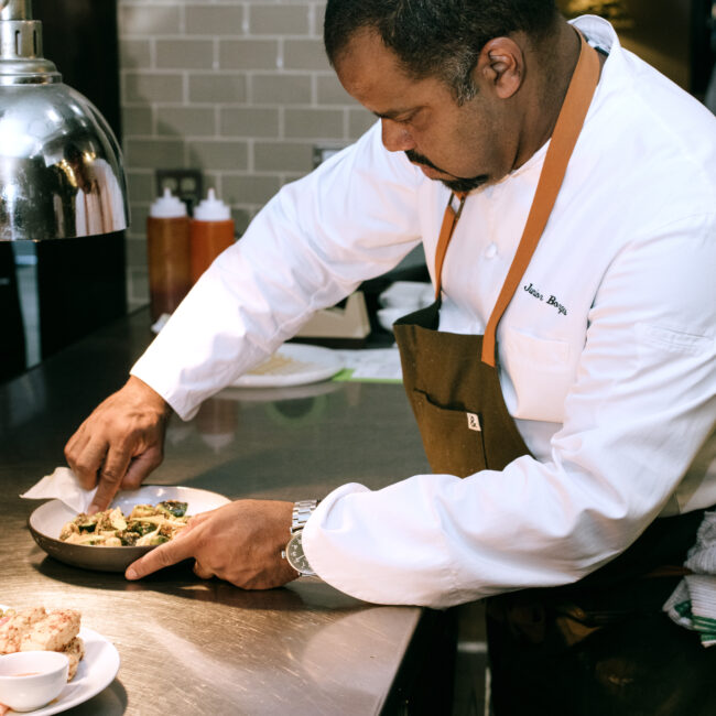 Chef Junior plating a dish at the kitchen pass at Nic + Junior’s, wearing a white coat and brown apron