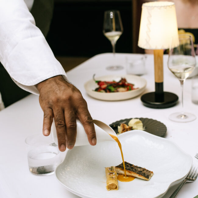 A server pours golden sauce tableside over a branzino fillet in the tasting room at Nic + Junior’s, with wine glasses and elegant plating in the background.