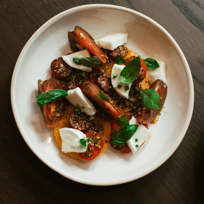 Heirloom tomato salad with fresh mozzarella, basil, and olive oil on a white plate at Nic + Junior’s